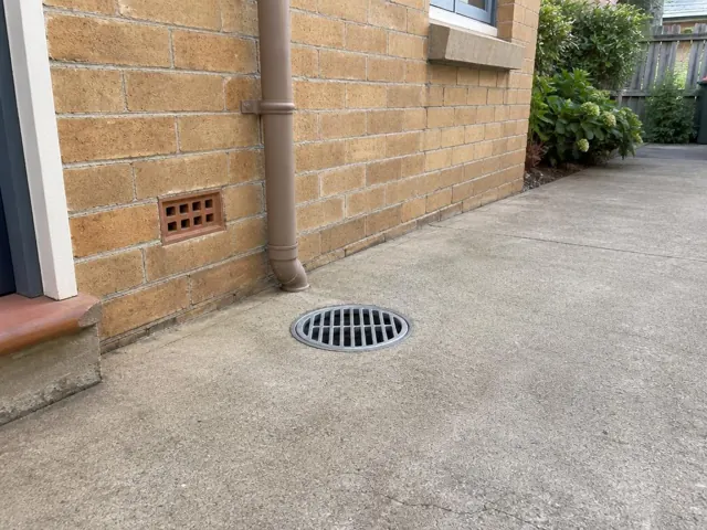 Outdoor concrete drain grate beside a brick house wall and downpipe with dry paving in Bondi